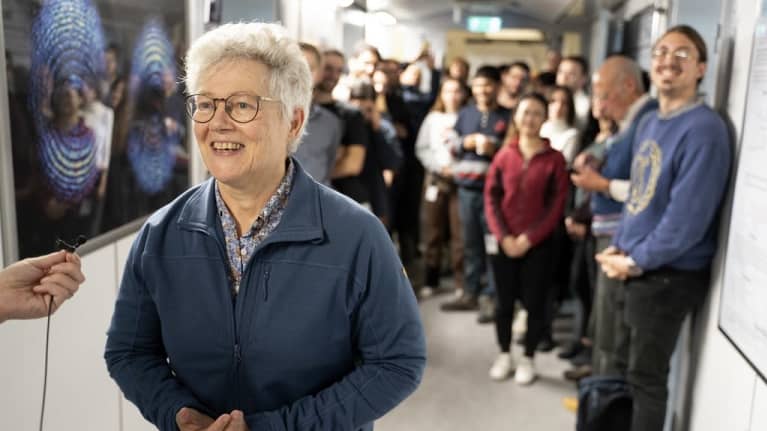 Hymyilevä Anne L'Huillier vastaamassa haastattelijan kysymykseen. Taustalla joukko iloisia opiskelijoita. 