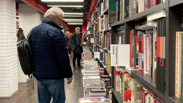 Pitkänomaisessa kirjakaupan salissa ihmiset tarkastelevat kirjoja hyllyn edessä. Etualalla on mies sinisessä talvitakissa reppu selässään. Hyllyssä näkyy selitysteksti: Kansalaissota.