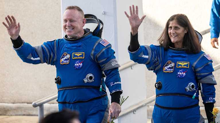 Kaksi sinisissä avaruuspuvuissa olevaa astronauttia vilkuttaa.