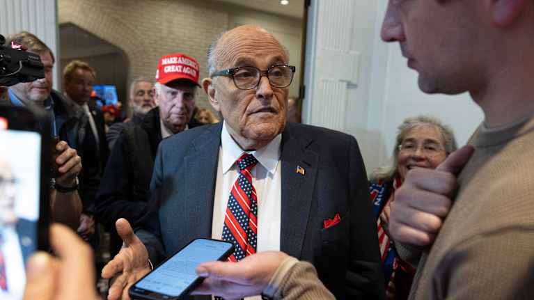 Rudy Giuliani puku päällä, puhuu älypuhelinta ojentavalle ihmiselle. Etualalla ihminen kuvaa Giuliania puhelimella. Taustalla ihmisiä. Yhdellä on päässä punainen lippalakki, jossa lukee Make America Great Again.
