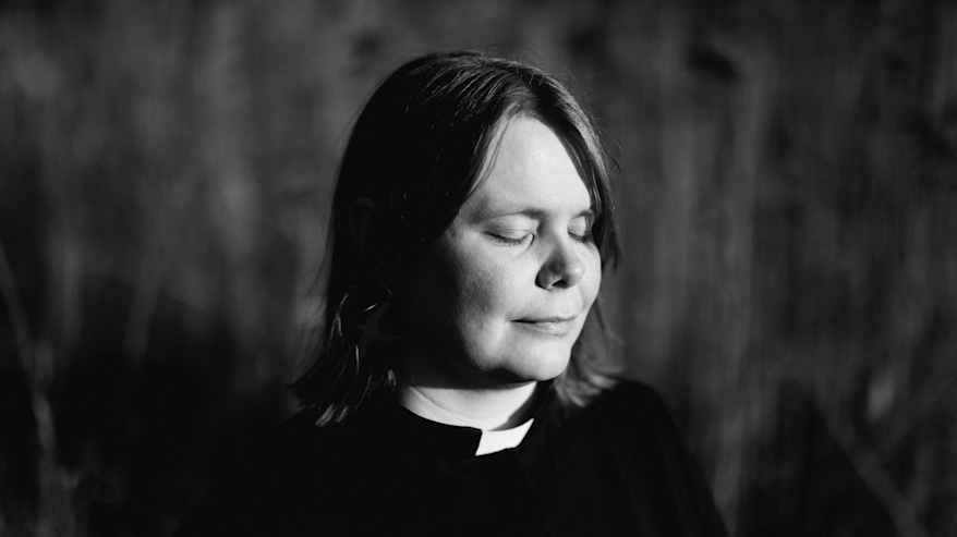Henkilö seisoo ulkona silmät suljettuina papin asussa.