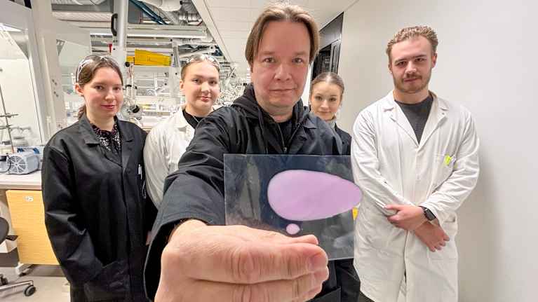 Mies katsoo suoraan kohti kameraa ja näyttää sille kädessään olevaa pientä näytelappua. Taustalla on neljä laboratoriotakkeihin pukeutunutta henkilöä, jotka hekin katsovat kohti kameraa.