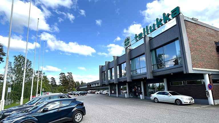 Punatiilinen  ja lasijulkisivuinen pitkä rakennus lähikuvassa. Rakennuksen eli Hotelli Tallukan vieressä on autoja rivissä parkissa. 
