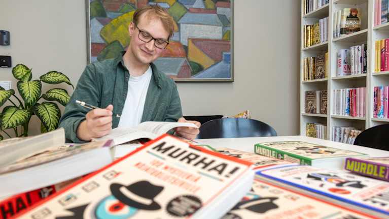 Henkilö istuu pöydän ääressä kirjapinon ympäröimänä, taustalla taulu ja kirjahylly.