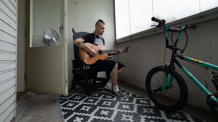 Nuori mies soittaa kitaraa parveella. Etualalla on polkupyörä.