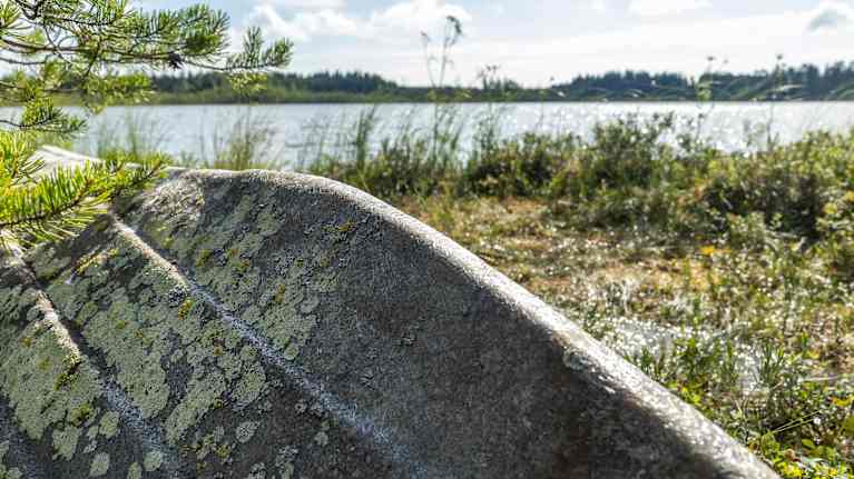 Näkymä järven rannalta, jossa maalipinnan menettänyt ja jäkälää kasvava lasikuituvene lepää ylösalaisin taustalla siintävänjärven rannalla.