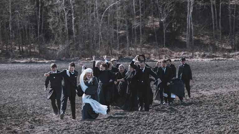 Iloinen joukkio kävelee pellolla. Kuvassa näyttelijöitä, joilla on 1800-1900-lukujen vaihteen juhlavaaatteet. Joukkion edessä kulkee hääpari.