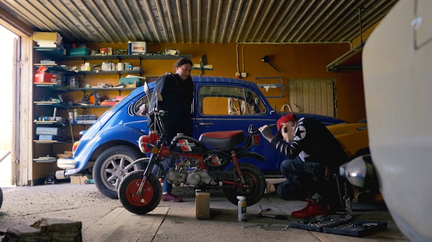 Nuori mies korjaa mopoa autotallissa. Taustalla on äiti ja kuplavolkkari.