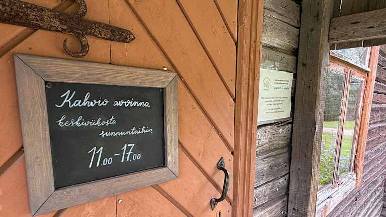 Vanhan puutalon ovessa on kyltti, joka kertoo kahvilan olevan avoinna keskiviikosta sunnuntaihin.