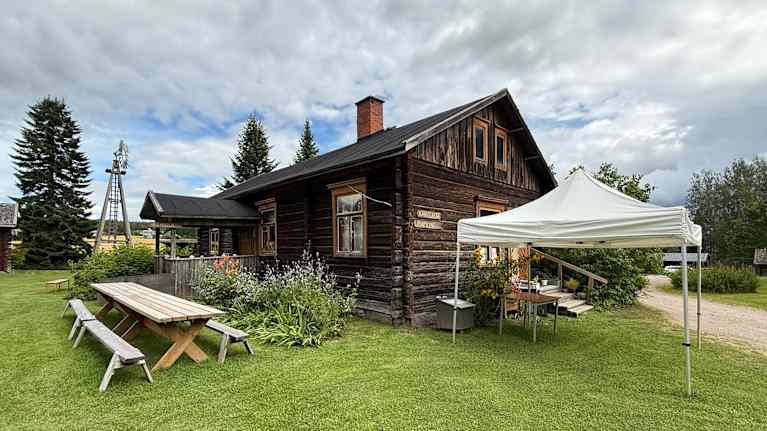 Vanha puurakennus keskellä kesäistä vihreää nurmikkoa, etualalla näkyy pöytiä ja penkkejä ja teltta. Rakennuksen ympärillä on kukkaistutuksia.