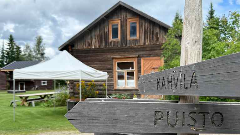 Taustalla näkyy vanha puutalo kesäisessä maalaismaisemassa, sen vieressä on valkoinen teltta. Kuvan etualalla on opasteviittoja.