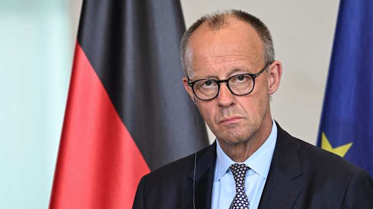 Saksan liittokansleri Friedrich Merz kuuntelee kokousta saksan ja Eu:n liput taustallaan.