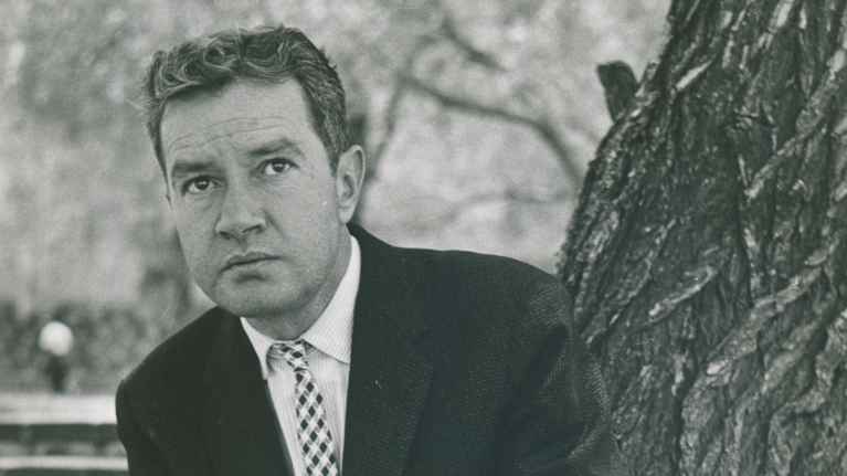 Mies pukeutuneena tyylikkääseen, 1950-luvun tyyliseen pukuun istuu ulkona puun vieressä.