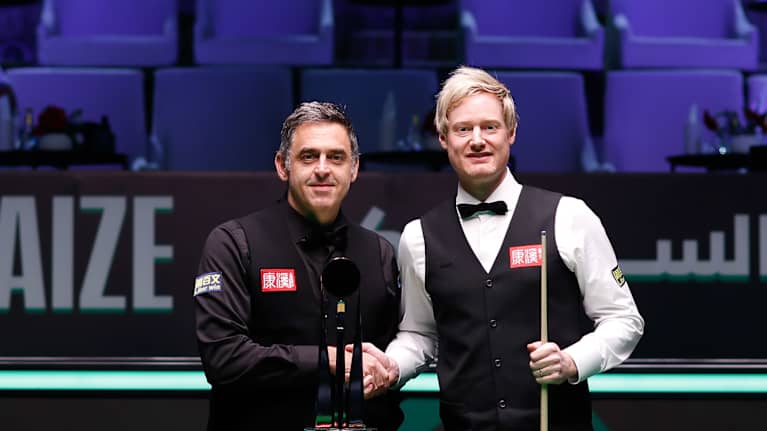 Ronnie O'Sullivan ja Neil Robertson kättelevät pelipöydän takana. Edessä voittopokaali.