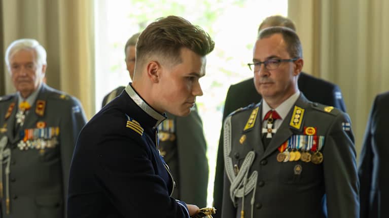 Sotilas vastaanottaa kunniamitalin seremoniallisessa tilaisuudessa. Kuvassa näkyy uniformuja ja kunniamerkkejä.