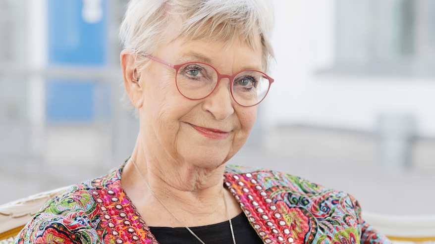 Tuula Karjalainen istuu sohvalla ja pitelee käsissään uusinta kirjaansa, katsoo sivusilmin kohti kameraa.