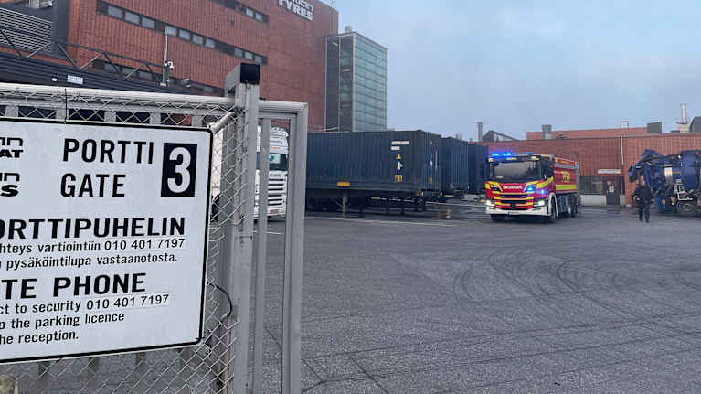 Nokian Renkaiden tehdasalueen portti 3, jossa pelastusajoneuvo on parkkeerattuna teollisuusrakennusten läheisyydessä. Taustalla näkyy kontteja ja savua.