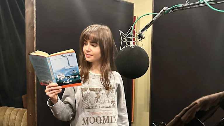Muumi-paitaan pukeutunut Lily Collins pitelee kirjaa studiossa.