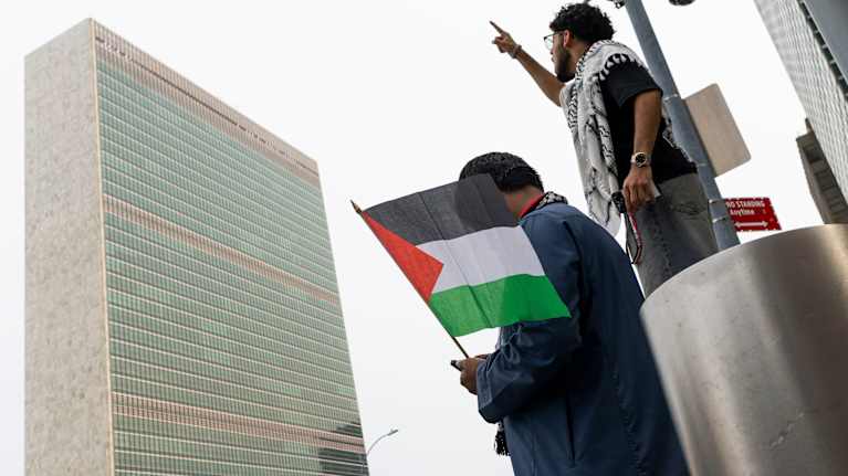 Kaksi miestä seisoo YK:n päämajan edessä, toisella Palestiinan lippua ja toisella palestiinalaishuivi.