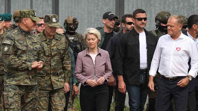 Ursula von der Leyen keskellä. Vasemmalla puolella sotilaspukuisia miehiä, oikealla Puolan pääministeri Donald Tusk ja ilmeisesti turvamiehiä.