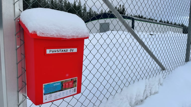 Metalliaidassa punainen postilaatikko, jossa lukee Puustako Oy. Verkkoaidan takana näkyy teollisuushalli.