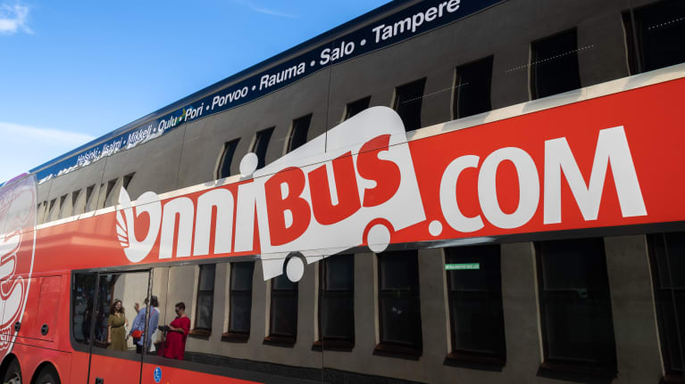 Onnibus linja-auton kylki. Linja-auton ikkunan heijastuksesta näkyy kyytiä odottavia ihmisiä aseman edessä.