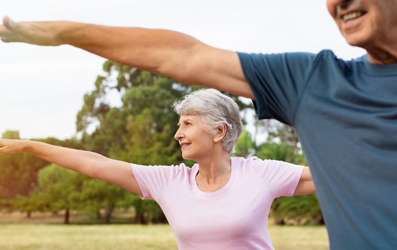 Kuvituskuva. Iäkäs nainen ja mies jumppaamassa puistossa. Ulkona on aurinkoinen kesäpäivä.