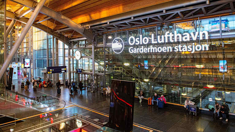Ihmisiä seisoo ja istuu odottamassa lentoasemalla. Seinällä kyltissä lukee "Oslo Lufthavn".