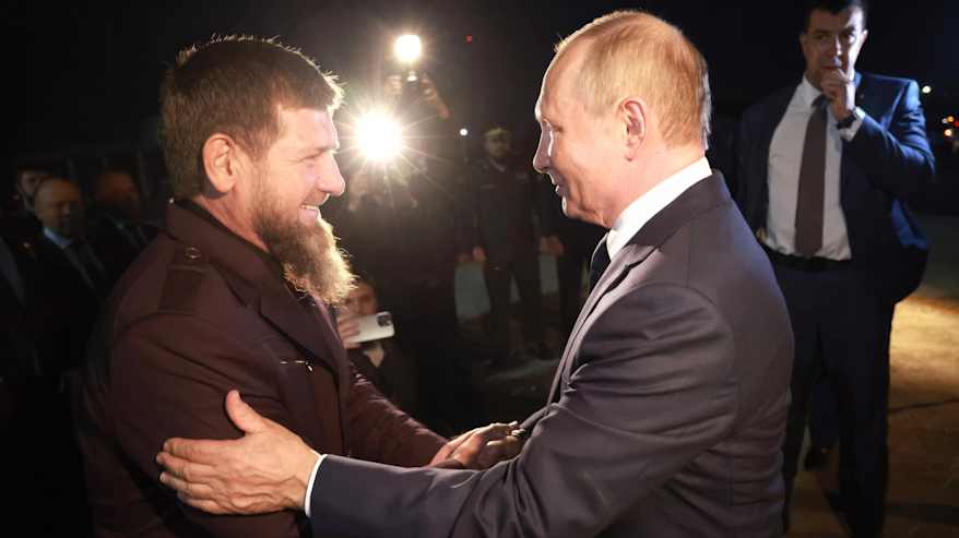Ramzan Kadyrov ja Vladimir Putin kättelevät hymyillen. Putin pitää vasenta kättään Kadyrovin käsivarrella. Putinilla on tummanharmaa puku, Kadyrovilla tumma asu. Taustalla näkyy kuvaaja, jonka kameran valo loistaa pimeydessä.