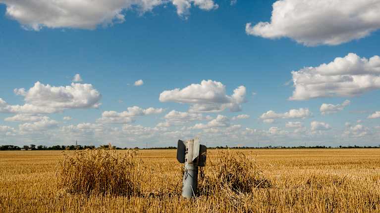 Keskellä peltoa on maassa törröttävä pommi, ja taustalla levittäytyy poutapilvinen sininen taivas.