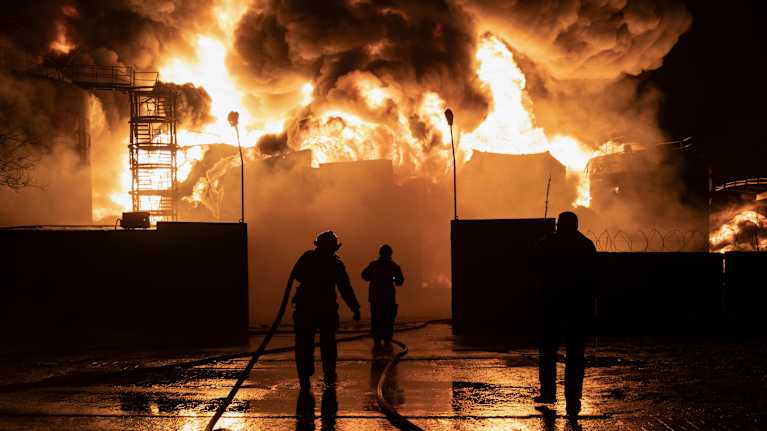 Palomiehet taistelevat kiivaasti valtavaa tulipaloa vastaan. Liekkimassat valtaavat taustan, kun tiimi pyrkii hallitsemaan tilannetta vesiletkujen avulla.