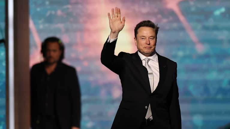 Elon Musk tervehtii yleisöä tapahtumassa. Hänellä on puku päällä. Taustalla seisoo mies, joka on kuvassa epätarkka.
