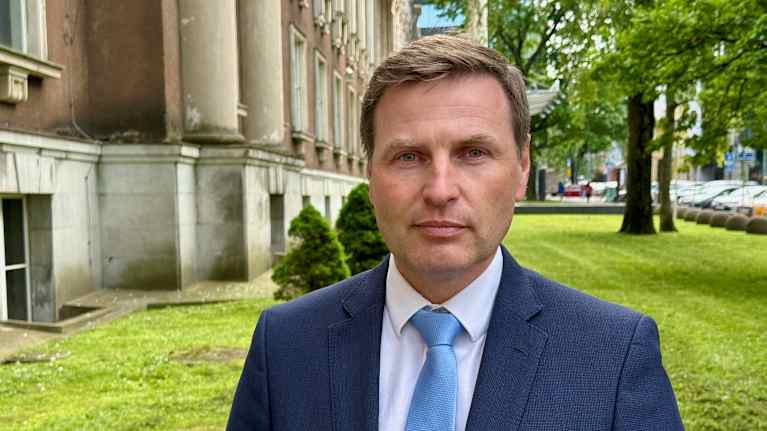 Mies puvussa, kauluspaidassa ja solmiossa seisoo vanhahkon kivitalon edessä nurmikolla ja katsoo suoraan kameraan.