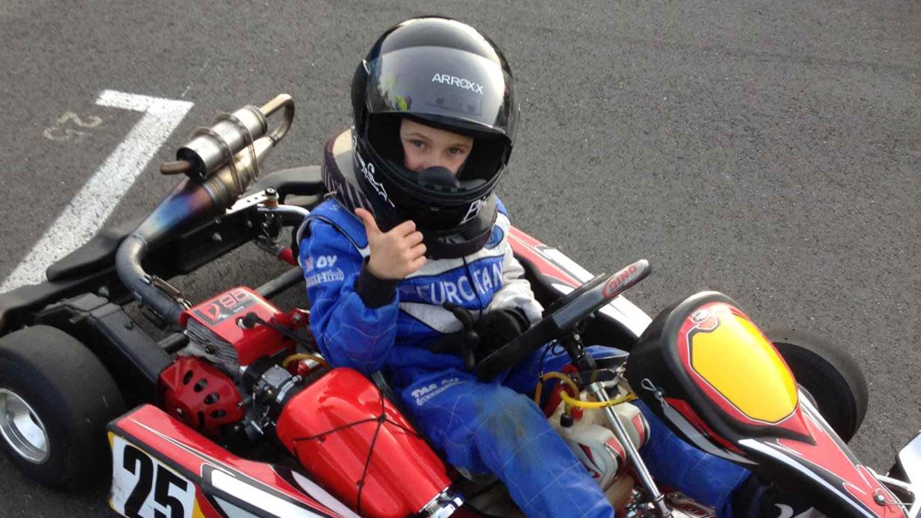 8-vuotias poika istuu karting-auton kuskin paikalla ja näyttää kameralle peukkua.