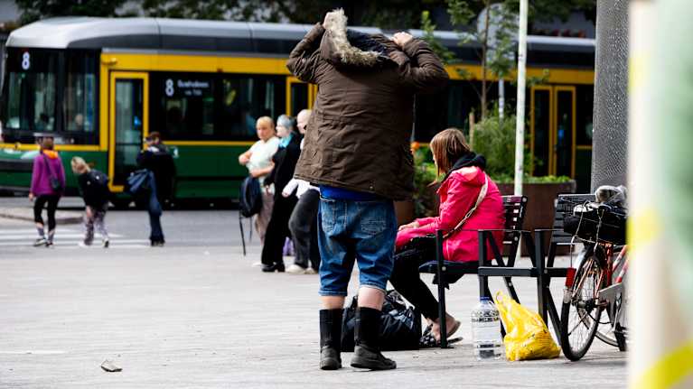Toppakenkiin ja farkkusortseihin pukeutunut mies pukee päälleen talvitakkia. Taustalla raitiovaunu.