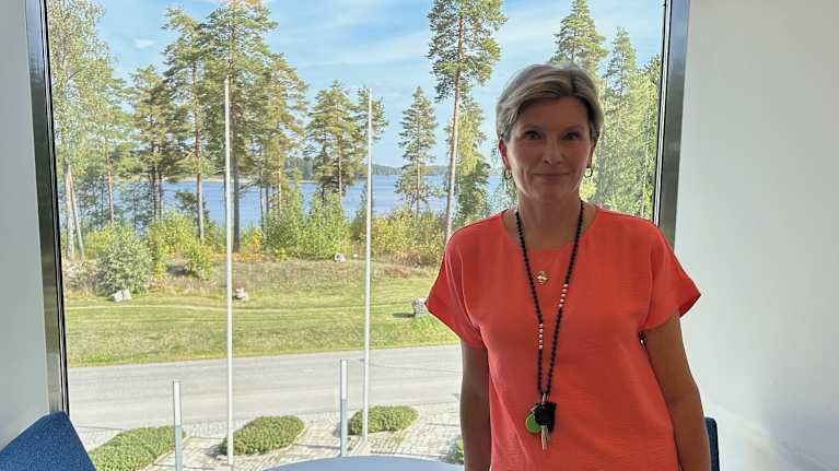 Nainen seisoo upeassa järvimaisemassa valoisassa huoneessa, pukeutuneena kirkkaan oranssiin t-paitaan.