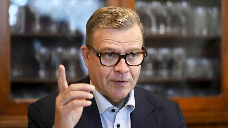 Petteri Orpo puhuu ja tekee käsillään eleitä puunvärisen kirjahyllyn edessä. 