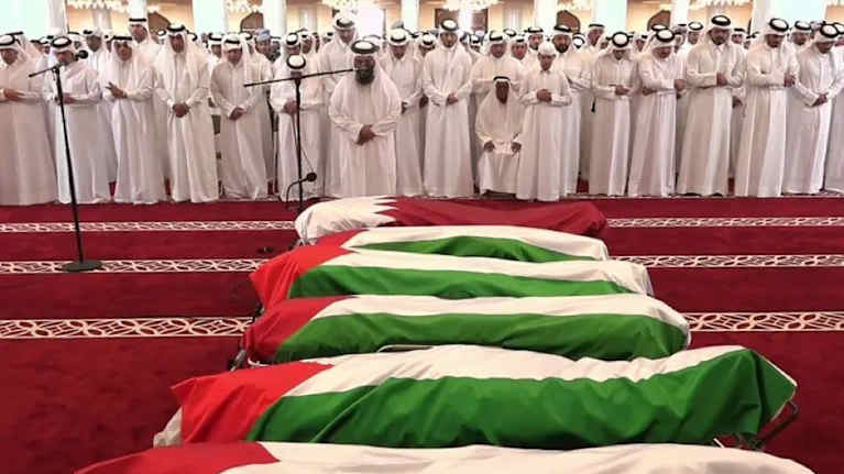 Valkoisiin kaapuihin ja päähineisiin (thawb ja ghutrah) pukeutuneita miehiä, kuuden arkun edessä. Arkut on peitetty Palestiinan ja Qatarin lipuilla.