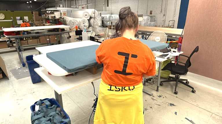 Kuvassa on oranssipaitainen nainen, joka ompelee kangasta kiinni levyyn Iskun tehtaalla Lahdessa. 