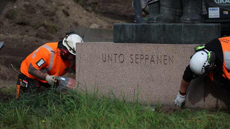 Huomiovaatteisiin pukeutunut mies pitää jonkinlaista konetta patsaan juurella, toinen koskee patsaan alustaa maantasalla.