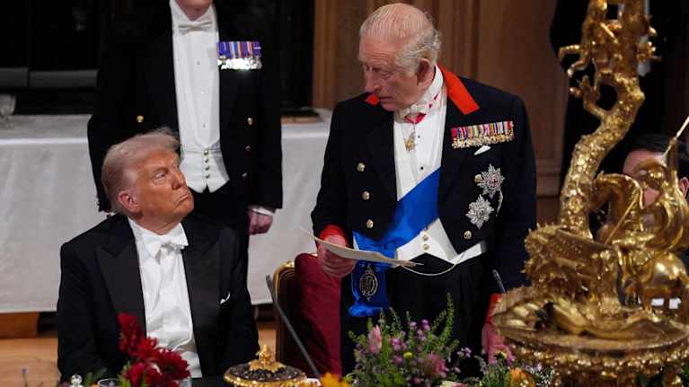 Donald Trump istuu juhlapöydän äärellä ja kuningas Charles puhuu seisaaltaan vierellä. Miehet katsovat toisiaan.