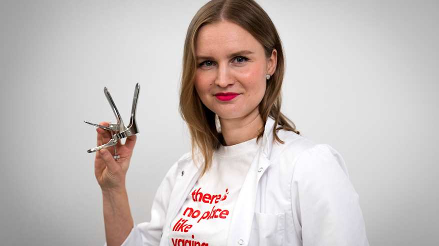 Kia Lampi seisoo lääkäritakki päällään seinän edessä ja pitää oikeassa kädessään gynekologista tutkimusvälinettä.