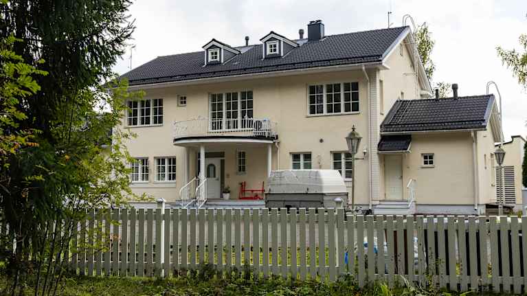 Suuri vaalea talo valkoisen aidan takana.