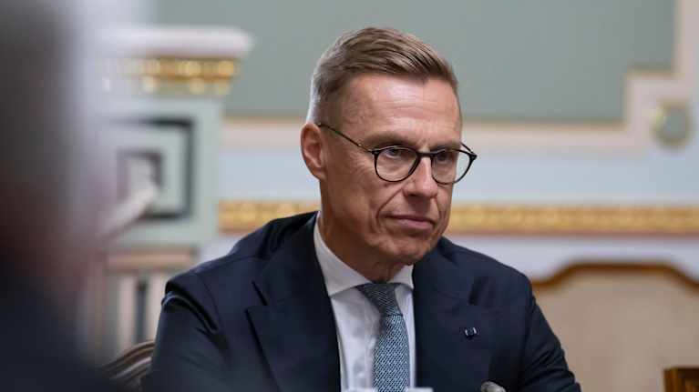 En närbild av Alexander Stubb i mörkblå kostym. Han sitter vid ett bord med en mikrofon och två vattenflaskor framför sig och ser allvarlig ut.