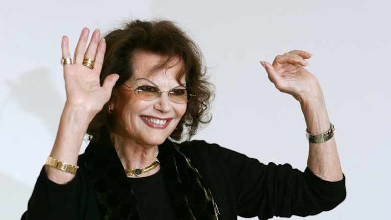 Tummiin pukeutunut vanhempi nainen hymyilee ja vilkuttaa valokuvaajille elokuvafestivaaleilla.