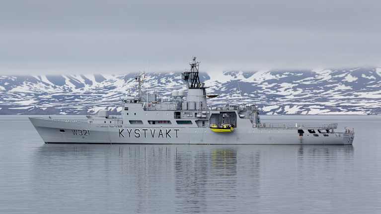 Norjan rannikkovartioston alus KV Senja lumisessa vuonomaisemassa, taustalla lumista rantaa ja matalalla roikkuva pilvipeite.