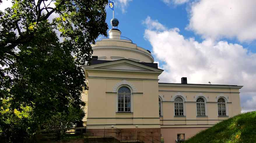 Uusklassinen vaalea rakennus, jossa matalahko pyöreä torniosuus, huipulla sininen pallo sekä Turun vaakunalippu, etualalla puun lehvästöä.