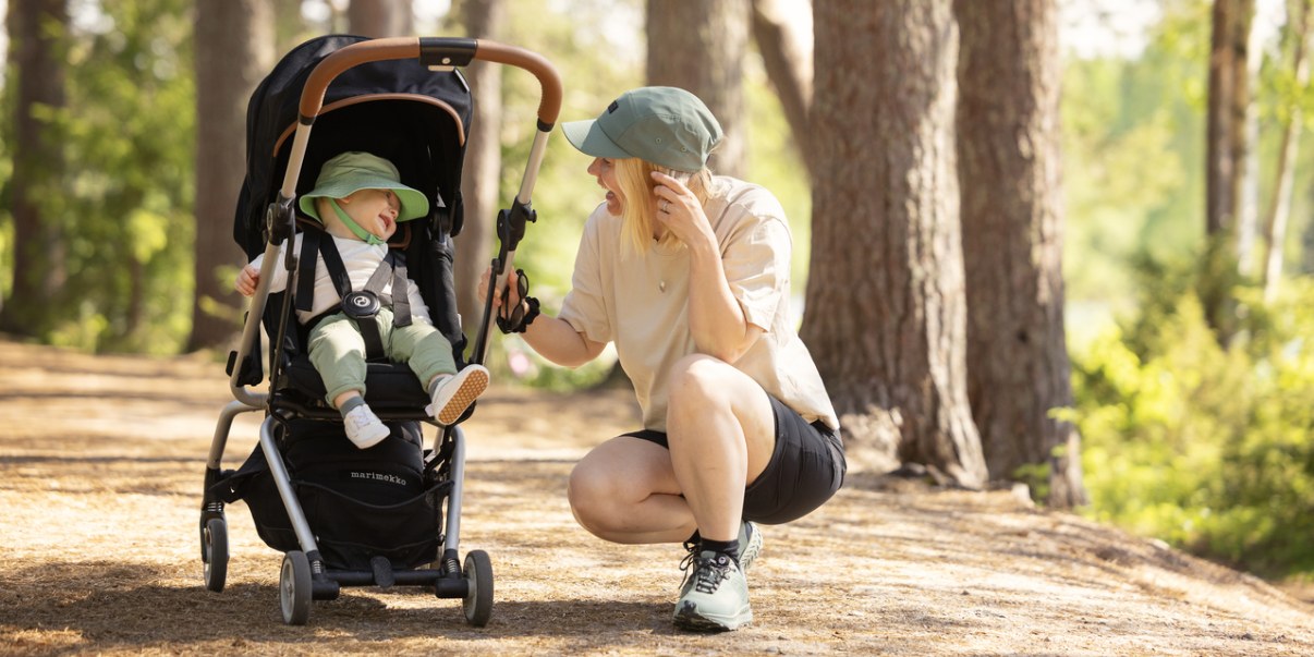 Metsäpolulla pieni lapsi rattaissa ja äiti kyykistyneenä rattaiden viereen. Äiti hymyilee lapselle.