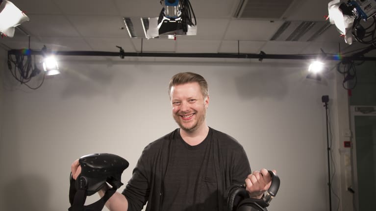 Mies poseeraa studiossa virtuaalilasit ja kuulokkeet kädessä.