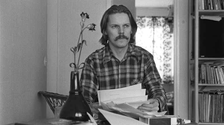 Arto Melleri istuu pöydän luona, paperinippu kädessä.